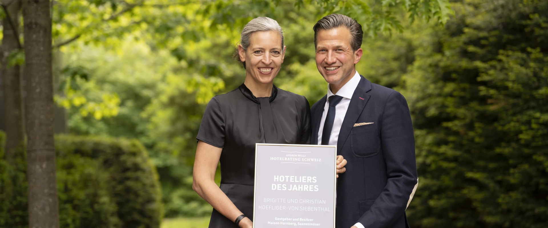 Zwei strahlende Hoteliers mit Auszeichungsurkunde vor grünem Naturhintergrund